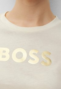 Camiseta de color crema con un prominente logotipo en dorado que dice "BOSS". La tela parece suave con una textura sutil y un clásico escote redondo.
