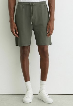 Mees, kes kannab oliivrohelisi tekstuuriga paelaga lühikesi pükse, valgeid sokke ja valgeid tossusid, seisab ühevärvse valge seina ääres.