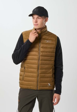 Jeune homme portant un gilet matelassé marron par-dessus une chemise noire à manches longues et une casquette noire, ajustant le col du gilet, debout devant un fond uni.