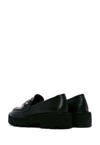 Mocassins à plateforme en cuir noir avec une semelle épaisse, comportant un ornement décoratif en argent à l'avant et une texture lisse.