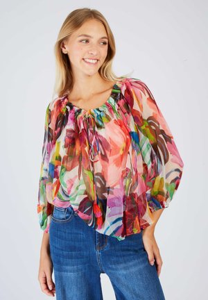 Young woman wearing a colorful floral sheer blouse with puffed sleeves and blue jeans, smiling and looking to the side.