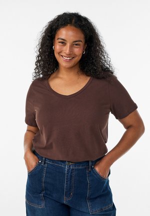 Kvinde med krøllet hår iført en brun V-hals T-shirt og blå jeans, smilende med hænderne i lommerne mod en ensfarvet baggrund.