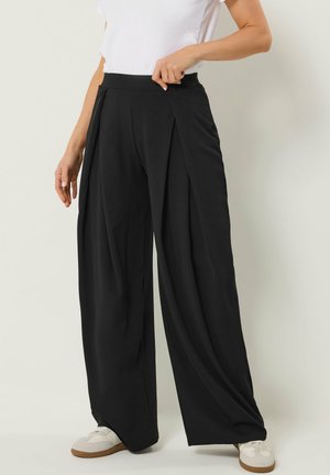 Donna che sistema pantaloni neri a vita alta e gamba larga abbinati a sneakers bianche e una maglietta bianca a maniche corte su uno sfondo semplice.