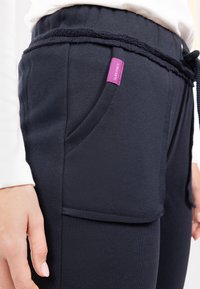 Navy zils sporta bikses ar auklu joslu, sānu kabatu ar šuvju detaļu un purpura zīmola etiķeti uz kabatas.