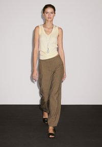 Una modella cammina con sicurezza su una passerella, indossando un top giallo e pantaloni larghi marroni, con una collana d'argento e sandali neri.