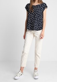 Donna che indossa una blusa a maniche corte blu navy con pois bianchi, pantaloni avorio e sneakers bianche con pois neri, in piedi contro uno sfondo semplice.