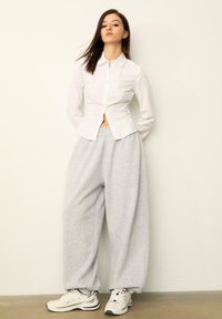 Weißes Hemd mit Knopfleiste und tailliertem Schnitt, kombiniert mit lockeren, grauen Jogginghosen und weißen Sneakern. Schlichtes Design und lässige Texturen.