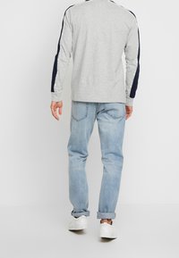 Grijze longsleeve shirt met marineblauwe schouderstrepen en witte accenten, gecombineerd met lichtblauwe jeans en witte sneakers. Over het geheel een gladde textuur.