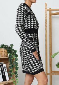Femme portant une robe cardigan à motif pied-de-poule noir et blanc avec des boutons, se tenant à côté d'une étagère en bois et de plantes.