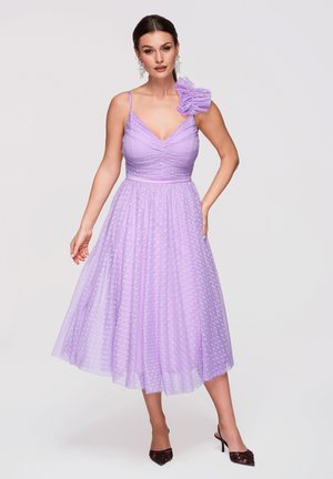 Donna che indossa un abito midi lavanda senza maniche con una sovrapposizione di tulle a pois e una grande balza su una spalla, abbinato a tacchi neri.