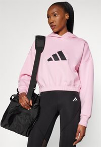 Rožinė trumpa džemperis su juodu Adidas logotipu, derinama su juodomis leginsomis, turinčiomis mažą baltą logotipą, ir juodu petiniu krepšiu.