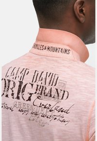 Lichtroze polo shirt met een zachte textuur, geborduurd kraag en bedrukte graphics met "CAMP DAVID" en "MOUNTAINS."