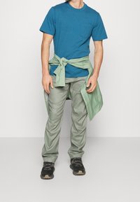 Tyrkysové bavlněné tričko s kapsou na hrudi, spárované se světle zelenými, mírně strukturovanými kalhotami. Černé tenisky dokončují outfit.