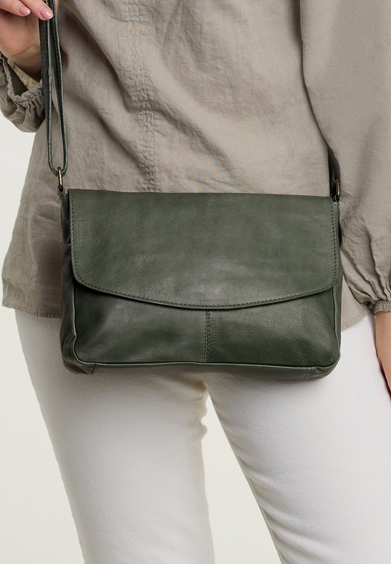Borsa a tracolla verde in pelle con chiusura a pattina, texture liscia e una sottile tracolla, tenuta a mano contro un outfit dai toni chiari.