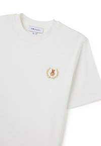 Camiseta blanca de manga corta con un pequeño oso de peluche bordado dentro de una corona de laurel en el lado izquierdo del pecho y un cuello redondo.