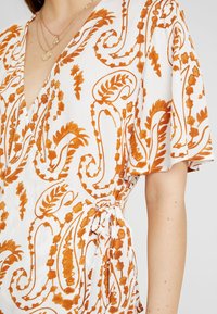 Chemisier blanc avec motif floral orange, présentant un design croisé, des manches courtes à volants et un détail de lien sur le côté. Tissu texturé.