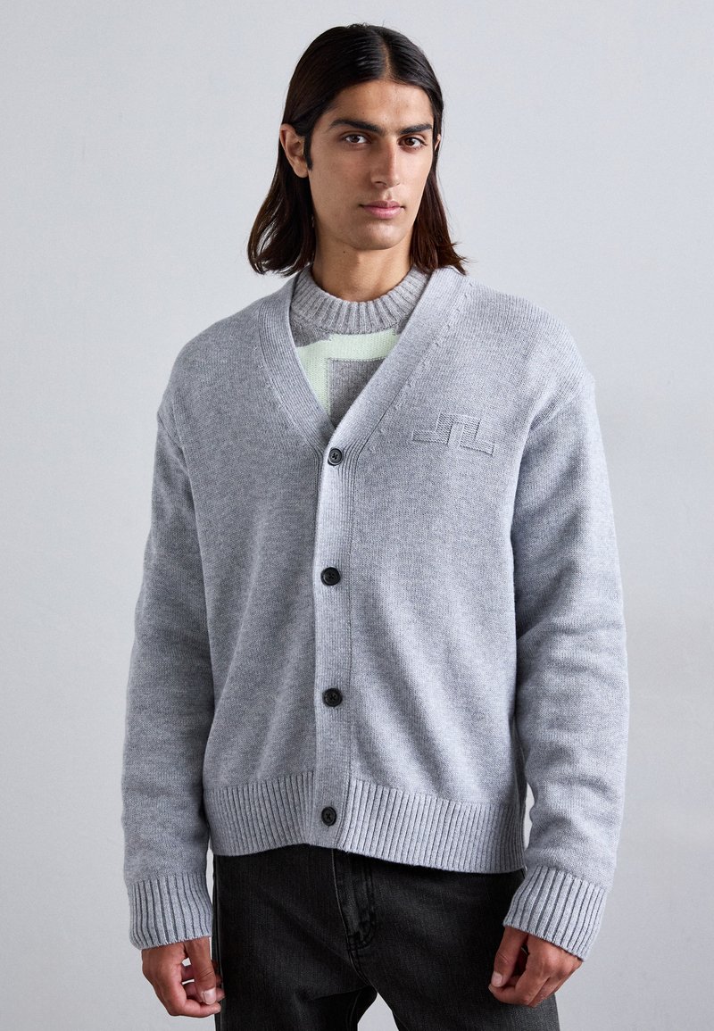 Cardigan grigio realizzato in morbido tessuto a maglia, con scollo a V, cinque bottoni, polsini a coste e un logo discreto sul petto. Indossato sopra un maglione a fantasia.