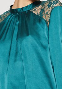 Blouse en satin bleu sarcelle avec un col montant, ornée de détails en dentelle sur les épaules. Texture lisse avec des plis créant un effet fluide.