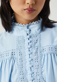 Lys blå bluse med dekorativ blondedetalj, knappelukking og rynket stoffdetaljering langs fronten og ermene.