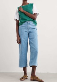 Green knitted vest over a white t-shirt, paired with high-waisted light blue wide-leg jeans and brown sandals with yellow straps.