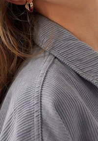 Jonge vrouw draagt een gouden gedraaide oorring en een grijze corduroy overhemd met zichtbare kraag en schoudernaden.