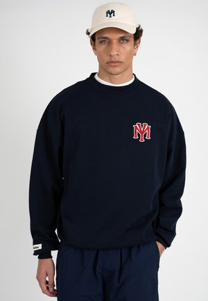 Joven con sudadera azul marino con iniciales rojas en el pecho y gorra blanca con iniciales azul marino, manos en los bolsillos, expresión neutra, fondo claro.