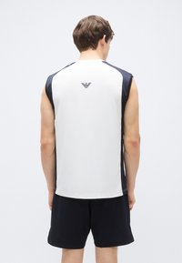 Top atlético sem mangas com parte da frente branca, painéis laterais azuis escuros e um logótipo nas costas. Tecido liso com um corte moderno. Usado com calções pretos.