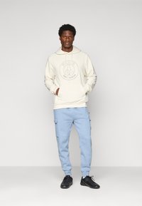 Sweatshirt com capuz creme com logótipo de Paris, bolso canguru; calças de fato de treino azul claro com bolsos laterais; ténis pretos. Textura suave, corte casual.