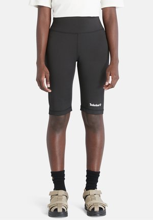 Osoba nosi czarne obcisłe spodenki do kolan z logo Timberland, czarne skarpetki, beżowe sandały na paski oraz biały krótki top, stojąc.