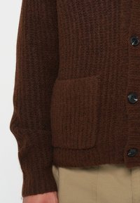 Brown knit cardigan with vertical ribbing, single chest pocket, black buttons, and textured fabric, featuring fitted cuffs at the sleeves.