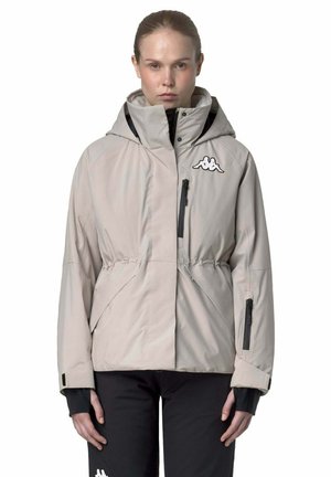 Donna che indossa una giacca invernale beige con cappuccio, zip nere e logo sul petto, abbinata a pantaloni neri con logo vicino al ginocchio.