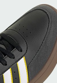 Crne atletske tenisice s kožnim i velur teksturama, s bijelim i žutim trakama, vezanjem na vezice i perforiranim detaljima na prstima.