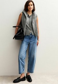 Vrouw in een grijze mouwloze gepersonaliseerde sweater, blauwe jeans, zwarte loafers, met een zwarte handtas, staande tegen een witte muur.