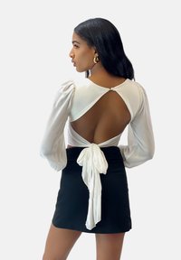 Haut blanc à manches longues avec des manches bouffantes et un dos ouvert, maintenu par un nœud. Associé à une mini jupe noire.