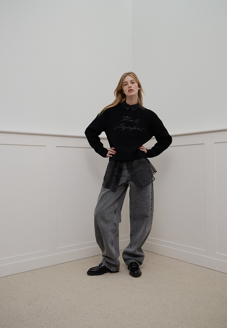 Un pull noir avec un texte imprimé, superposé sur une chemise à col grise, associé à un pantalon gris ample et des chaussures noires sur un fond uni.