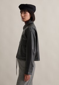 Schwarze Lederjacke mit einer taillierten Gestaltung, Taschen vorne und Kordelzug am Saum. Kombiniert mit einem grauen Pinstripe-Rock und einer schwarzen Baskenmütze.