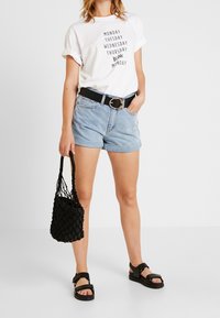 Bílý tričko s černým textem, světle modré džíny, černý pásek s kulatou sponou, černá tkaná taška a černé sandály.