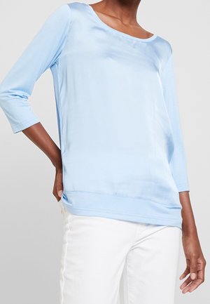 Femme portant un haut à manches longues en satin bleu clair avec un décolleté bateau et un pantalon blanc, posant avec une main sur la hanche devant un fond uni.