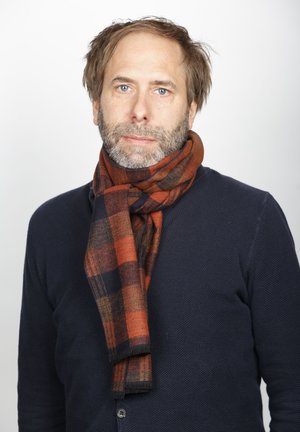 Erwachsener Mann mit hellbraunen Haaren und Bart, trägt einen marineblauen Pullover und einen orange-schwarz karierten Schal, schaut neutral nach vorne.