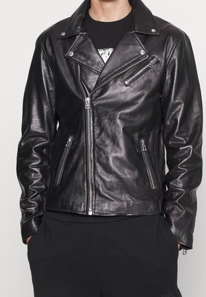 Homme portant une veste de motard en cuir noire avec des fermetures éclair argentées et une chemise noire, mains dans les poches sur un fond uni.