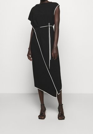 Mujer con un vestido midi negro con ribete blanco y dobladillo asimétrico, combinado con sandalias negras de tacón alto con tiras.