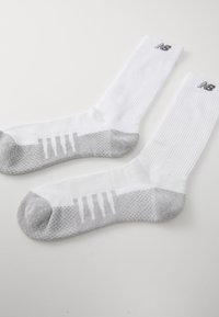 Calcetines deportivos blancos con textura acanalada, secciones de talón y punta en gris, y rayas blancas verticales en la parte gris; logo de New Balance en la parte superior.