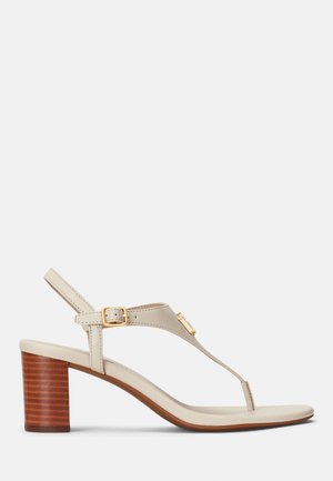 Sandale en cuir beige avec un petit talon en bois empilé, design à bride en T, boucle dorée et bride de cheville sur fond blanc.