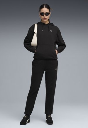 Kobieta w czarnej bluzie z kapturem i spodniach, czarnych trampkach, ciemnych okularach przeciwsłonecznych, trzymająca białą torebkę na ramię, stojąca na szarym tle.