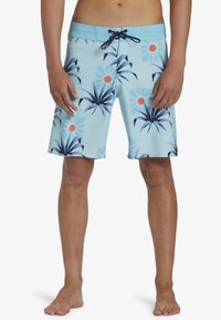 Costume da bagno blu con coulisse nera e bianca, caratterizzato da foglie di palma blu scuro e motivi grafici di sole rosso, indossato da una persona in piedi.