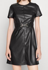 Svart faux läderklänning med korta ärmar, rund halsringning, bälte i midjan och silverfärgade detaljer, med en figurnära överdel och en utsvängd kjol.