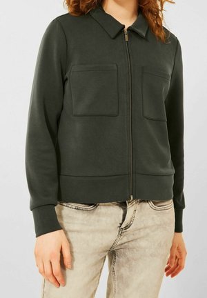 Vrouw die een donkergroen jack met rits draagt, met twee borstzakken en licht beige gewassen jeans, staand tegen een effen achtergrond.