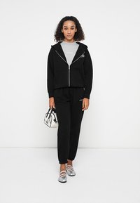 Felpa nera con zip e fodera interna grigia, abbinata a pantaloni della tuta neri. Gli accessori includono una piccola borsa bianca e mocassini argentati lucidi.