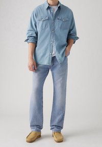 Camisa de mezclilla azul claro con botones en el frente y bolsillos en el pecho, combinada con jeans azul claro y zapatillas estilo mocasín beige.