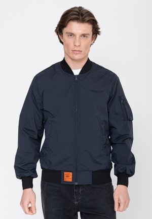 Man draagt een navy bomberjack met geribbelde manchetten en zoom, een ritssluiting aan de voorkant en een klein oranje patchje bij de taille, tegen een witte achtergrond.